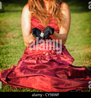 eine Frau in einem roten Kleid sitzt auf einer Wiese, eine Feder in ihren Händen hält Stockfoto