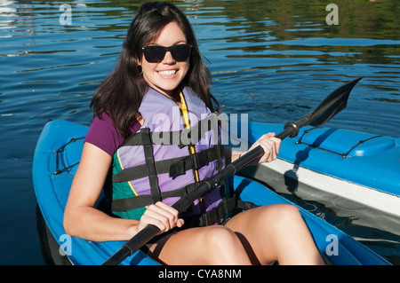 Eine lächelnde junge Brünette Frau trägt eine Schwimmweste sitzt im Kajak mit einem Paddel bereit, ihr Abenteuer beginnen. Stockfoto