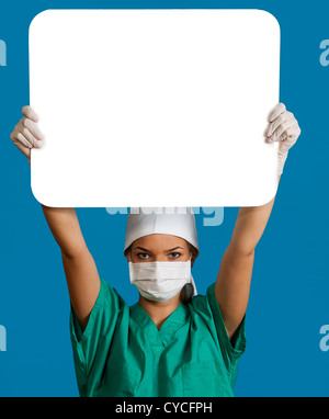 Eine junge Ärztin in einer Maske hält eine leere Bill-Board über den Kopf vor einem blauen Hintergrund. Stockfoto