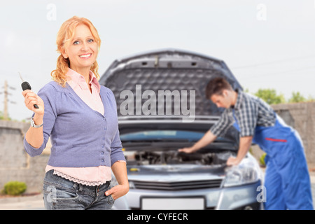 Lächelnd unvorsichtige weiblich eine Taste in der Hintergrund-Mechaniker prüft ihr Auto Stockfoto