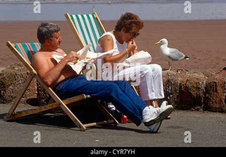 Ein Urlaub-paar sitzen in Liegestühlen, eingewickelt in Papier, die traditionelle Lebensweise für Essen Fish And Chips Chips zu genießen. Stockfoto