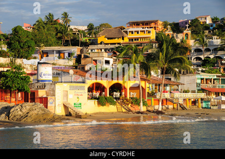 Puerto Escondido, Mexiko, Oaxaca, Playa Principal. Farbenfrohen Gebäuden unter Kokospalmen säumen den Strand. Stockfoto