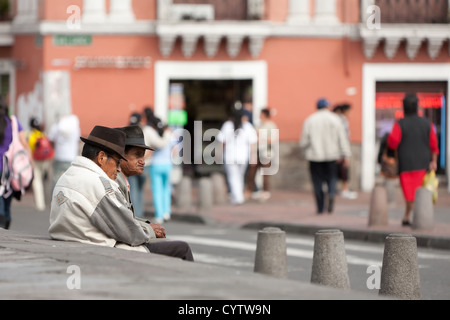 Zwei ältere Männer mit Fedora-Hüten sitzen nebeneinander auf einem Betonrand in einer städtischen Straßenlage. Der Mann im Vordergrund trägt eine helle Jacke mit einem Patch auf der Schulter und dunkel Stockfoto