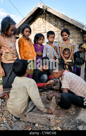 LUANG NAMTHA, Laos — Ein kleiner Junge in einem Dorf in der Provinz Luang Namtha im Norden von Laos, schneidet sorgfältig eine hölzerne Spinnplatte für das traditionelle Tujlub-Spiel. Er integriert eine Metallspitze von einem Schirmrücken, um einen haltbaren Punkt für dieses handgefertigte Spielzeug zu schaffen, das in einem konkurrenzfähigen Spiel mit Schnur verwendet wird, das bei lokalen Kindern beliebt ist. Stockfoto