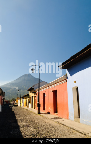 Kopfsteinpflasterstraße in Richtung Volcan de Agua Antigua Guatemala | ANTIGUA GUATEMALA, Guatemala — Schwaches Morgenlicht streift eine Kopfsteinpflasterstraße, die mit ockerfarbenen, gelben und hellblauen Kolonialfassaden gesäumt ist, wobei der Kegel des Volcán de Agua am fernen Ende in Antigua Guatemala aufragt. Der Lahar des Stratovulkans aus dem Jahr 1541 zerstörte die frühere Kolonialhauptstadt an Almolonga und veranlasste ihre Verlegung ins Tal von Panchoy im Jahr 1543. Dicke, niedrige Mauern entlang der Straße spiegeln Jahrhunderte des Bauens für seismische Überlebensfähigkeit wider. Stockfoto