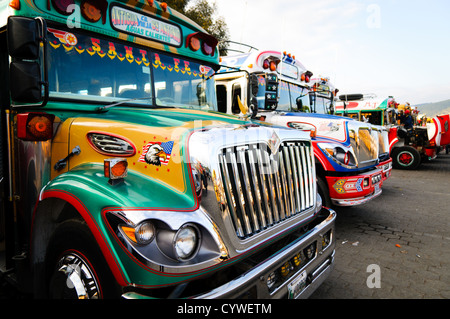 Chicken Buses Antigua Guatemala | ANTIGUA GUATEMALA, Guatemala – Eine Reihe bunt bemalter Hühnerbusse, umfunktionierte amerikanische Schulbusse, die mit Chromgittern und handschriftenbeschrifteten Zieltafeln ausgestattet sind, wartet auf einer Straße in Antigua Guatemala. Diese Camionetas sind nach wie vor das Rückgrat des lokalen Verkehrs durch Guatemala, dekoriert von ihren Besitzern in Stilen, die zu einer anerkannten Volkskunst-Tradition geworden sind. Stockfoto