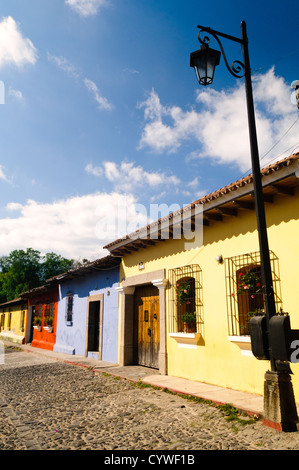 Kopfsteinpflaster-Häuser Antigua Guatemala | ANTIGUA GUATEMALA, Guatemala — Eine Reihe von Kolonialhäusern entlang einer Kopfsteinpflasterstraße zeigt die Mischung aus gelben, blauen und roten Fassaden, die typisch für das historische Viertel der Stadt ist, mit eisernen Fensterläden, Topfpflanzen und einer schmiedeeisernen Straßenlaterne. Der UNESCO-geschützte Kern von Antigua bewahrt dieses niedrige koloniale Layout weitgehend unverändert seit der spanischen Herrschaft. Stockfoto