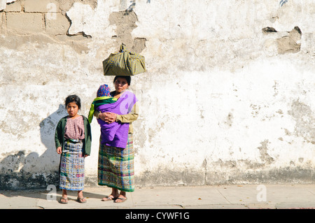 Guatemaltekische Frau mit Kindern Antigua Guatemala // ANTIGUA GUATEMALA, Guatemala — Eine guatemaltekische Frau posiert mit ihrer Tochter und ihrem Baby auf einer Straße. Sie trägt ihr Baby in einer traditionellen Tragetasche und einem großen Bündel auf dem Kopf, während ihre Tochter neben ihr steht. Beide sind in traditioneller Kleidung gekleidet und stehen an einer verwitterten Wand, die typisch für die historischen Straßen von Antigua ist. Stockfoto