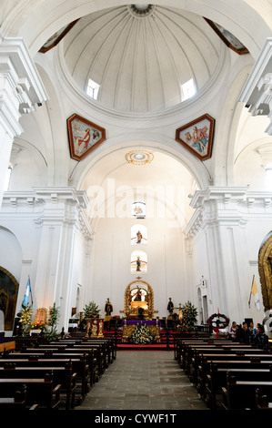Iglesia de San Francisco Altar Interior Antigua Guatemala // ANTIGUA GUATEMALA, Guatemala — Inneres und Altar der Iglesia de San Francisco. Die große Inneneinrichtung verfügt über eine hohe gewölbte Decke und eine markante Kuppel, die durch natürliches Licht aus den oberen Fenstern beleuchtet wird. Verzierte goldene Details zieren den Altar und stehen im Kontrast zu den überwiegend weißen Wänden und dunklen Holzbänken der Kirche. Stockfoto