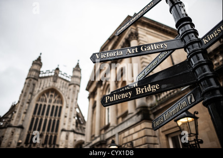 Bath Abbey Wegweiser Bath Vereinigtes Königreich | BATH, Vereinigtes Königreich — Eine Gruppe von gusseisernen Wegweisern zeigt Richtung Pulteney Bridge und der Victoria Art Gallery, während der Turm der Westfassade der Bath Abbey hinter ihnen im kompakten historischen Kern der Stadt aufragt. Die Abtei, die ab 1499 unter Bischof Oliver King neu gebaut wurde, ist das Herzstück einer UNESCO-Weltkulturerbestadt, in der die wichtigsten Sehenswürdigkeiten in Gehweite voneinander liegen. Stockfoto