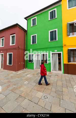 Eine Frau in einem roten Mantel vor bunt bemalten Häusern in Burano, in diesem Fall Erbsen-Grün, Zitronengelb und roten Ocker Stockfoto