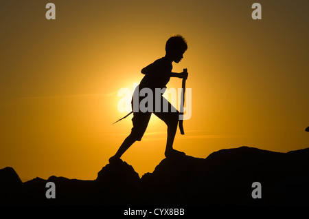 Eine Freude junge spielt auf den Felsen von einem Pier mit Holzstöcken während der Dämmerung. Stockfoto