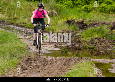 Eine Frau auf ihr Mountainbike durch eine schlammige Pfütze auf einem Feldweg in der Nähe von Edmundbyers in Northumberland. Stockfoto