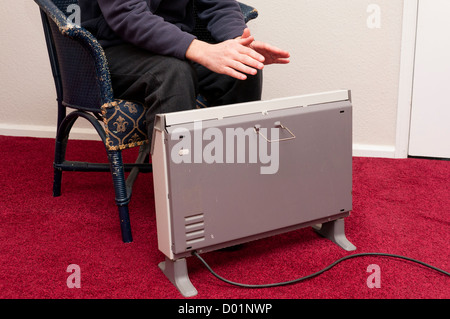 Energiearmut UK. Mann in Fleece-Jacke sitzt neben alten Elektroheizung. Stockfoto