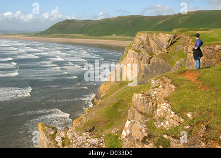 Frau steht auf einer Klippe mit Blick auf die Wellen und Strand, Rhossili Bucht, Halbinsel Gower, Wales, UK Stockfoto