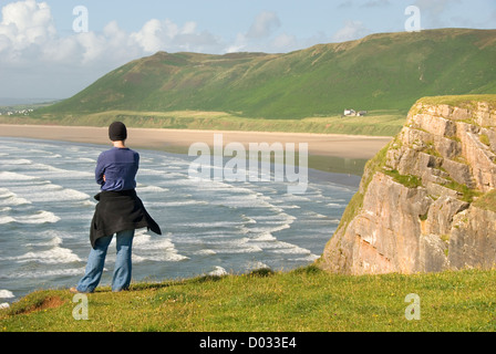 Frau steht auf einer Klippe mit Blick auf die Wellen und Strand, Rhossili Bucht, Halbinsel Gower, Wales, UK Stockfoto