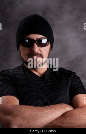 Stolz auf harter Kerl in schwarz. Starke Mann im schwarzen Hemd, Sonnenbrille und Zipfelmütze mit muskulösen Arme verschränkt, gerichtete Kamera Stockfoto