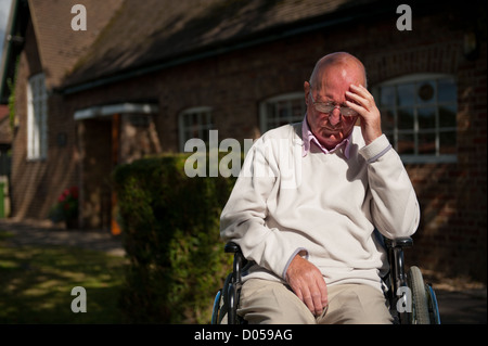 Älterer Mann sitzt im Rollstuhl mit seinem Kopf in der Hand.  Er ist im Freien vor seinem Haus/Pflege zu Hause. Stockfoto