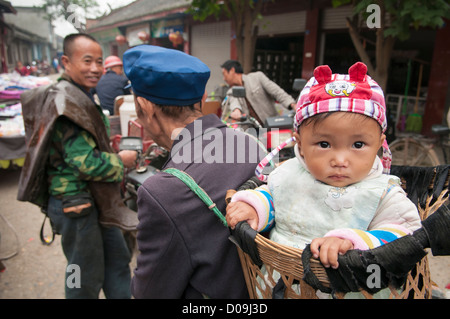 Baby Junge im Korb Großvater an belebten Markttag im Dorf am Stadtrand von Chengdu, Provinz Sichuan, China erfolgt durch Stockfoto