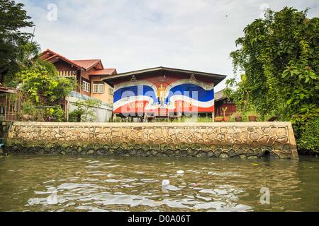 17. November 2012 - bemalt Bangkok, Thailand - traditionelle Gehäuse an einem Kanal im Abschnitt "Thonburi" von Bangkok, mit eine thailändische Flagge und eine Hommage an Bhumibol Adulyadej, der König von Thailand. Bangkok war früher bekannt als das "Venedig des Ostens '' aufgrund der Anzahl der Wasserstraßen, die Kreuz und quer die Stadt gekreuzt. Nun die meisten der Wasserstraßen ausgefüllt aber Boote und Schiffe nach wie vor eine wichtige Rolle im täglichen Leben in Bangkok. Tausende von Menschen pendeln täglich auf dem Chao Phraya Express Boote und Schnellboote, die ply Khlong Saen Saeb oder verwenden Boote auf den Kanälen auf die Thonbur zu umgehen Stockfoto