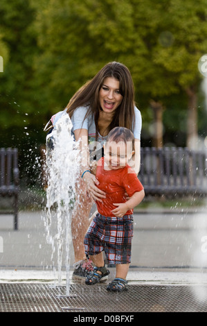 Junge Mutter erreichen für Kleinkind spielt in Brunnen im Waterfront Park in Louisville, Kentucky Stockfoto