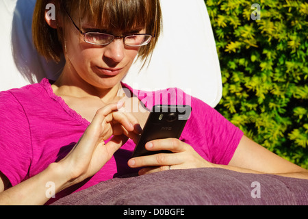 Eine Nahaufnahme von einer jungen Frau, überprüfen ihr Mobiltelefon während der Sitzung in einem sonnigen Garten. Stockfoto