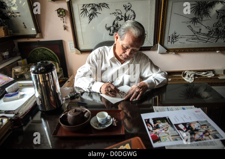 Kinh Van Le Hue Seidenstickerei-Künstler schreibt am Schreibtisch Hue Vietnam | HUE, Vietnam — Der lokale Künstler Kinh Van Le, ein Meister der traditionellen Hue-Seidenstickerei, schreibt an seinem Schreibtisch in seinem Geschäft, während Beispiele seiner Arbeit an der Wand hinter ihm ausgestellt sind. Hue gilt weithin als das führende Zentrum der Seidenstickerei in Vietnam, eine Handwerkstradition, die eng mit der langen Geschichte der Stadt als kaiserliche Hauptstadt der Nguyen-Dynastie verbunden ist. Die königliche Förderung der Bildenden Künste und Textilien durch die Stadt hat die Stickerei zu einer hohen Kunstform erhoben, und erfahrene Praktizierende wie Le setzen diese Tradition bis heute fort. Stockfoto