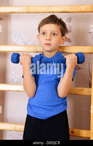 Der junge im Vorschulalter hebt Hanteln neben einer Sprossenwand Stockfoto