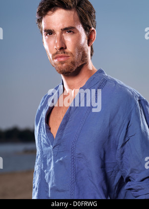 Mode-Porträt eines jungen Mannes trägt ein blaues Hemd gegen blauen Himmel am Strand Stockfoto