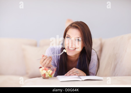 Eine attraktive junge Frau Obstsalat Essen und lesen auf der Couch liegend Stockfoto