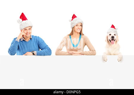 Ehepaar und Hund mit Weihnachtsmann-Mützen posiert hinter eine Blindplatte lächelnd Stockfoto