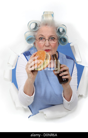 Ältere Frau mit ihrem Haar in Rollen einen Hamburger Essen und Bier zu trinken Stockfoto