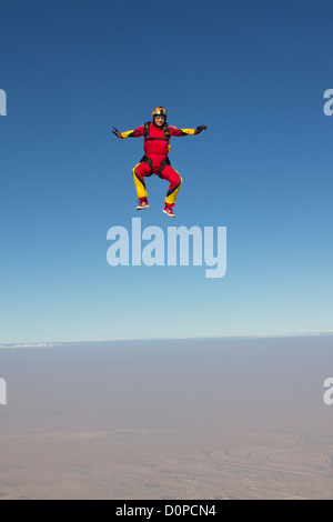 Fallschirmspringer-Mädchen in einer Stand-up-Position fällt frei durch den blauen Himmel über ein trockenes Wüstengebiet mit einem Lächeln auf ihr Gesicht. Stockfoto