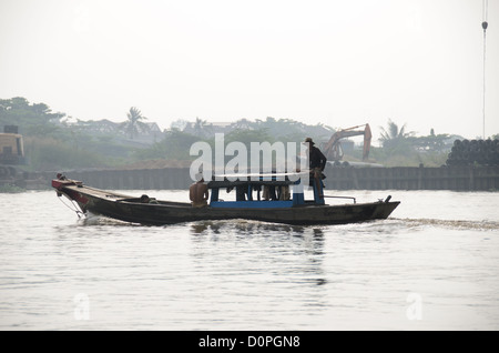 Traditionelles Holzboot Saigon River Ho-Chi-Minh-STADT Vietnam / HO-CHI-MINH-STADT, Vietnam — die Männer fahren mit einem traditionellen Holzboot entlang des Saigon River, vorbei an einem verschwommenen Flussufer, das mit Stapeln und schweren Baumaschinen gesäumt ist. Der rote Bug des Schiffes zeigt ein gemaltes „Boot Eye“ (*mat thuyen*), ein jahrhundertealter vietnamesischer Brauch, der Wassergeister abwehren und das Schiff sicher führen soll. Diese historische Flusskultur wird zunehmend parallel zu massiven städtischen Landgewinnungsprojekten betrieben, die die Küste der Stadt verändern. Stockfoto
