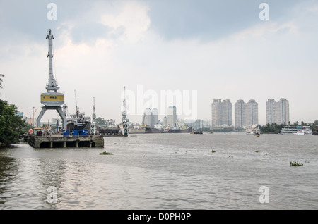 Industriekrane am Saigon-Fluss Moderne Skyline Ho-Chi-Minh-Stadt Vietnam | HO-CHI-MINH-STADT, Vietnam — Portalkrane flankieren den in Haiphong registrierten Frachter Thanh Hai 58 am Saigon-Fluss, während die Wohnhochhäuser der Stadt stromabwärts aufragen. Der Fahrwasser des Flusses, der für Tiefgänge von etwa 30 Fuß (9 m) schiffbar ist, ermöglicht es Schiffen, fast 50 Meilen (80 km) landeinwärts vom Südchinesischen Meer zu fahren, und historisch gesehen verbindet er die nördlichen und südlichen Häfen Vietnams. Stockfoto