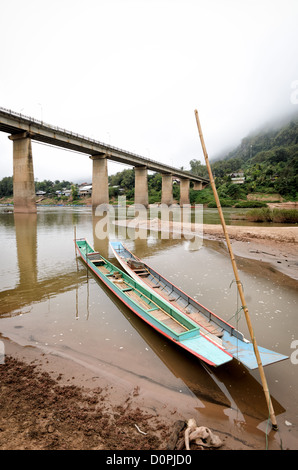 NONG KHIAW, Laos - Holzboote sind gefesselt Pole an den sandigen Ufern des Flusses Nam Ou (Organisationseinheit) in Nong Khiaw in Nord Laos zum Bambus. In der Mitte des Rahmens ist die hohe Brücke über den Fluss. Hinter dem werden Gebäude am Wasser. Nebel Verkleidung die steilen, felsigen Karste und Felsen in der Umgebung. Stockfoto