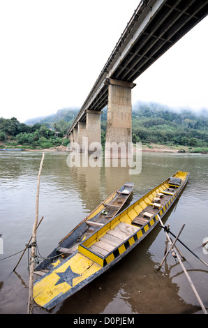 NONG KHIAW, Laos - Holz- und oben offenen Boote an einem Pfahl gebunden an den Ufern des Flusses Nam Ou (Organisationseinheit) direkt unter der hohen konkrete Brücke überspannt den Fluss in Nong Khiaw im Norden von Laos. Stockfoto