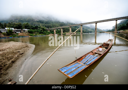 Langschwanz-Holzboot Nam Ou Fluss Brücke Nong Khiaw Laos | NONG KHIAW, Laos — Ein langes Holzboot liegt unter der Betonbrücke, die die beiden Hälften von Nong Khiaw über den Nam Ou Fluss verbindet, nebelverhangene Kalksteinklippen erheben sich dahinter. Die Brücke verbindet das Westufer der Stadt mit der Ostufer-Siedlung Ban Sop Houn, und der Nam Ou bleibt für kleine Boote bis hinauf nach Hatsa, nahe Phongsaly, schiffbar. Stockfoto