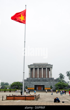 HANOI, Vietnam – Eine vietnamesische Flagge fliegt vor dem Ho Chi Minh Mausoleum, während Touristen auf dem Platz vor dem Hotel herumwandern. Das Ho-Chi-Minh-Mausoleum ist ein großes Denkmal in der Innenstadt von Hanoi, umgeben vom Ba Dinh-Platz. Es beherbergt den einbalsamierten Körper des ehemaligen vietnamesischen Führers und Gründungspräsidenten Ho Chi Minh. Stockfoto