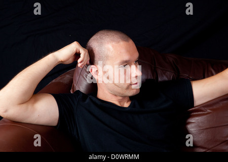 Mann mit enger geschnittenen Haaren an der Ecke der Couch zu sitzen und mit seinem Kopf auf seine Hand nach rechts schauend Stockfoto