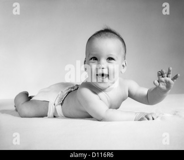 1960ER JAHREN LÄCHELN BABY TRAGEN STOFFWINDEL LIEGEND AUF BAUCH BETRACHTEN KAMERAARM ANGEHOBEN Stockfoto