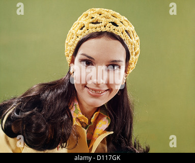 1970ER JAHRE PORTRAIT LÄCHELNDE JUNGE LANGE HAARE BRÜNETTE FRAU MIT GELBEN HÄKELN MÜTZE Stockfoto