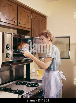 1960ER JAHREN FRAU HAUSFRAU IN SCHÜRZE OFEN BACKEN KOCHEN BRATEN IN KÜCHE Stockfoto