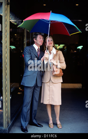 1970ER JAHREN PROFESSIONELLE MANN FRAU PAAR UNTER REGEN REGENSCHIRM Stockfoto
