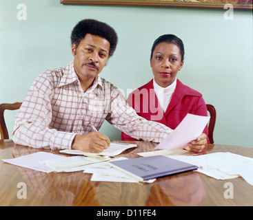 MITTE DER 1970ER JAHRE ALTER AFRIKANISCHER AMERIKANISCHER MANN FRAU PAAR SITZEN TISCH GEHEN ÜBER HAUSHALTSKONTEN BLICK IN DIE KAMERA Stockfoto