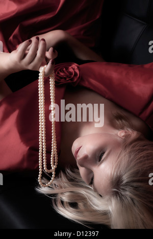 eine blonde Frau ist auf einem schwarzen Sofa liegend hält eine Perlenkette Stockfoto