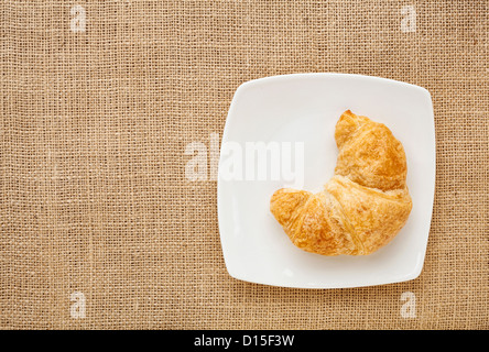Croissant-Rolle auf einen weißen Porzellanteller gegen Sackleinen Leinwand Brett Stockfoto