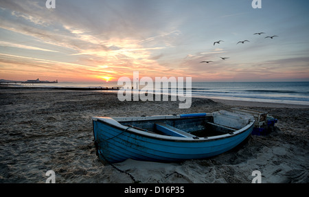 Türkis-Blau Fischerboot am Strand bei Sonnenaufgang mit Bournemouth Pier in der Ferne. Stockfoto