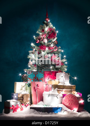 geschmückter Weihnachtsbaum mit roten Verzierungen und glänzend weiße Lichter, umgeben von bunten Geschenke mit Schnee bedeckt, vor blauem Hintergrund mit Farbverlauf Licht Stockfoto