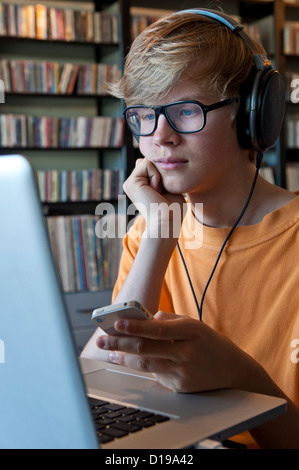 Nachdenklich Teenager Schuljunge mit Kopfhörern mit Apple iPhone Smartphone und Laptop-Computer mit Plattensammlung in b/g Stockfoto